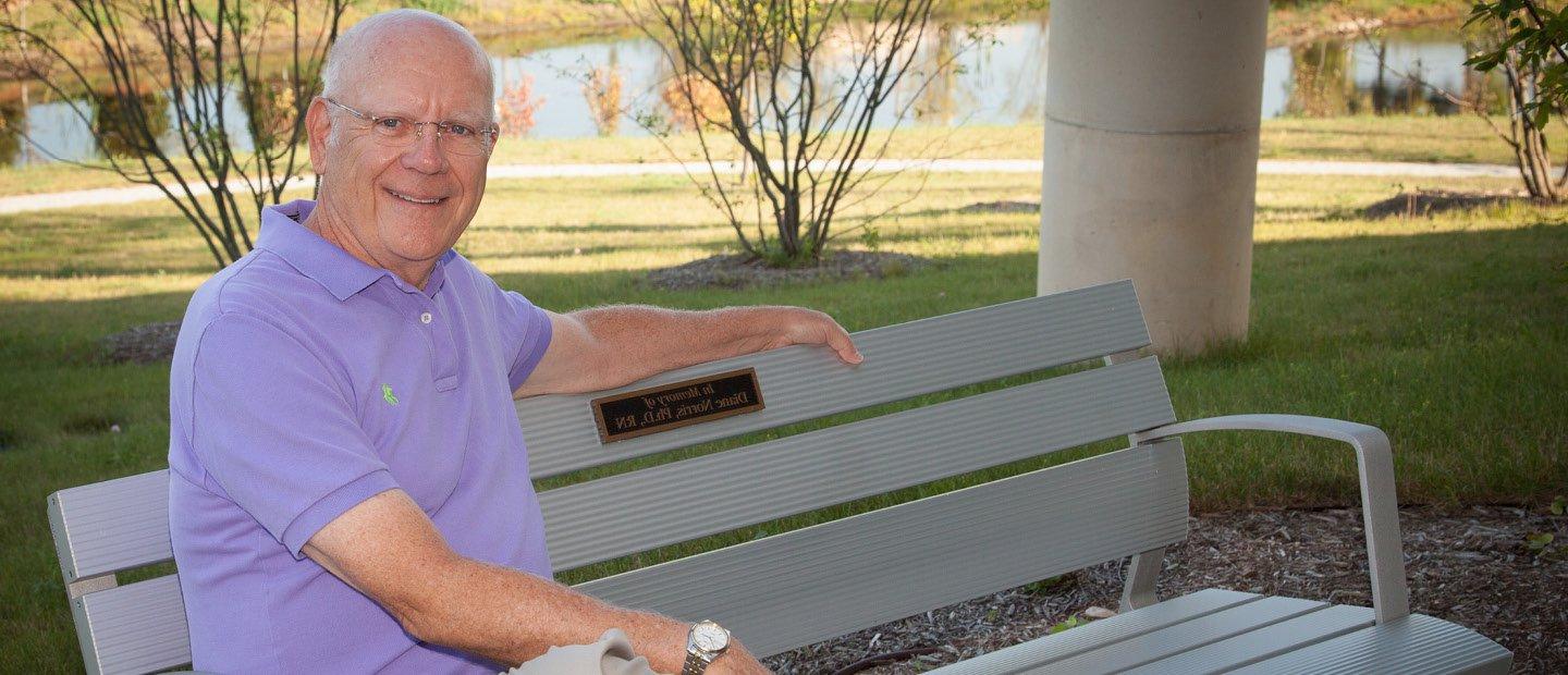 man sitting on a bench with a plaque that says "In Memory of Diane Norris, Ph.D., RN"