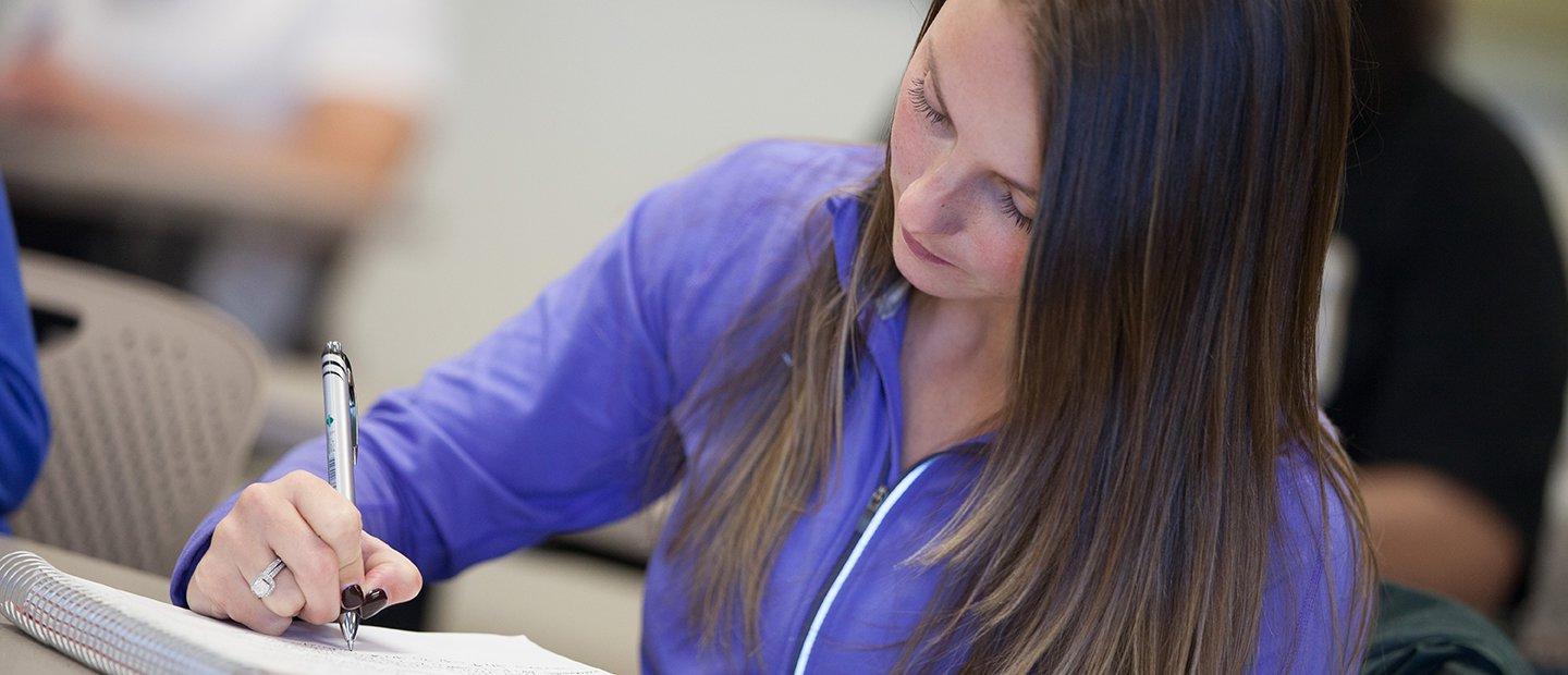 woman in a purple shirt seated at a desk, writing in a notebook